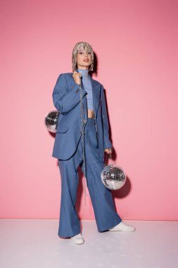 Full length of stylish young woman in jewelry headwear holding disco balls on pink background 