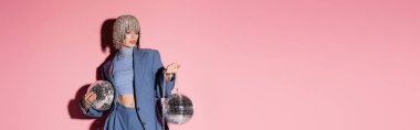 Trendy woman in luxury headwear and suit holding mirror balls on pink background, banner 