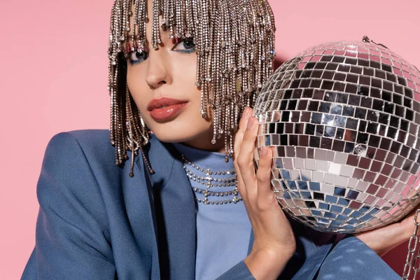 Stylish young woman in jewelry headwear holding mirror ball and looking at camera on pink background 