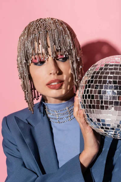 Trendy young woman in jewelry headwear looking at disco ball on pink background 