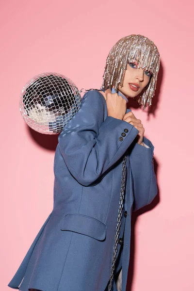 Trendy young woman in jacket and luxury headwear holding disco ball holding on pink 