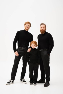 full length of adult bearded men and happy redhead boy standing with hands in pockets and looking at camera on grey background clipart