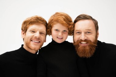 cheerful red haired boy with bearded grandfather and dad in black turtlenecks smiling at camera isolated on grey clipart