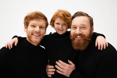 smiling redhead kid hugging bearded grandpa and dad wearing black turtlenecks isolated on grey clipart
