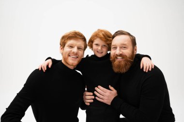 joyful red haired kid embracing dad and bearded grandfather in black sweaters isolated on grey clipart
