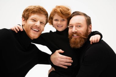 joyful redhead boy with father and bearded granddad embracing while looking at camera isolated on grey clipart