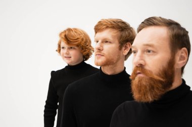 smiling redhead boy in black sweater looking away near father and bearded grandpa on blurred foreground isolated on grey clipart