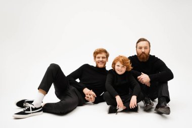 redhead boy sitting with crossed legs near joyful dad and bearded grandfather on light grey background clipart