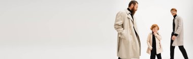 smiling redhead kid in trench coat posing with hand in pocket near father and granddad on light grey background, banner clipart