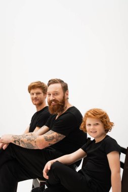 happy redhead boy sitting near bearded tattooed granddad and father isolated on grey clipart