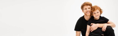 Siyah tişörtlü neşeli adam kızıl saçlı oğlanı kucaklıyor ve gri pankartta izole edilmiş kameraya bakıyor.