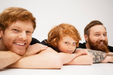 joyful man with redhead son and bearded tattooed dad lying with crossed arms and smiling at camera on light grey background clipart
