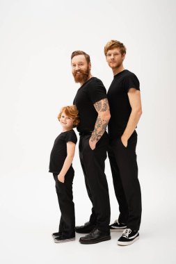 Uzun boylu, sakallı, kızıl saçlı, oğlu ve torunu olan açık gri arka planda siyah pantolon ceplerinde poz veren bir adam.