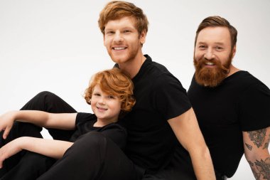 joyful red haired kid with father and bearded grandpa in black t-shirts looking at camera isolated on grey clipart