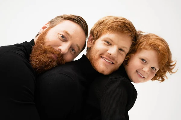 happy and bearded men with redhead boy smiling at camera while leaning on each other isolated on grey