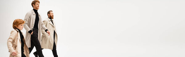 happy redhead boy and bearded men in stylish trench coats walking isolated on grey, banner