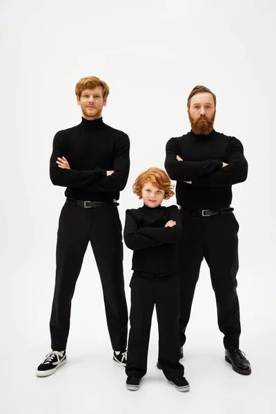 full length of bearded man with redhead son and smiling grandson posing with crossed arms on light grey background