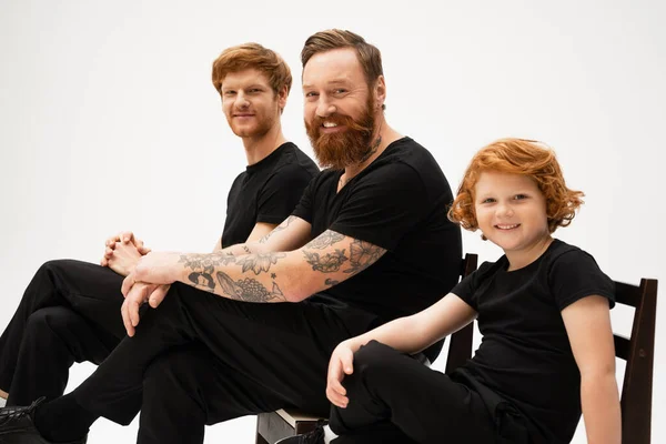 bearded tattooed man with redhead son and grandson smiling at camera while sitting isolated on grey