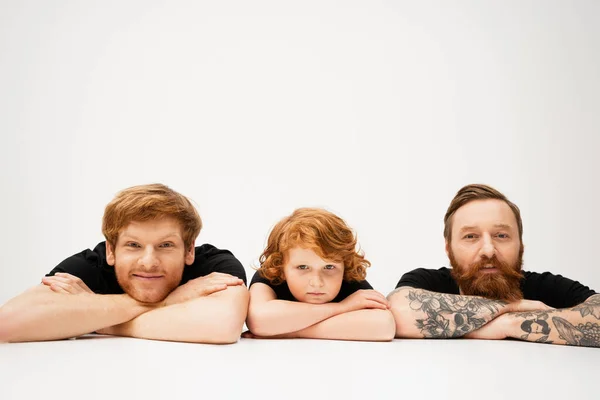 cheerful man with redhead son and bearded tattooed father lying with crossed arms on light grey background