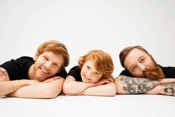 bearded tattooed man with redhead grandson and happy son smiling at camera while lying on light grey background