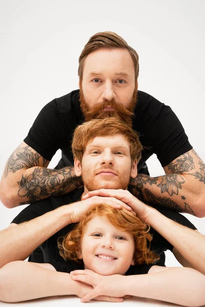 family portrait of bearded tattooed man with red haired son and grandson on light grey background