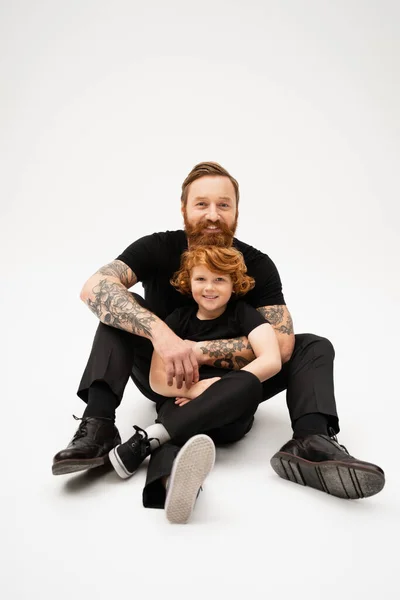 full length of tattooed man with red haired grandson sitting in black clothes on light grey background