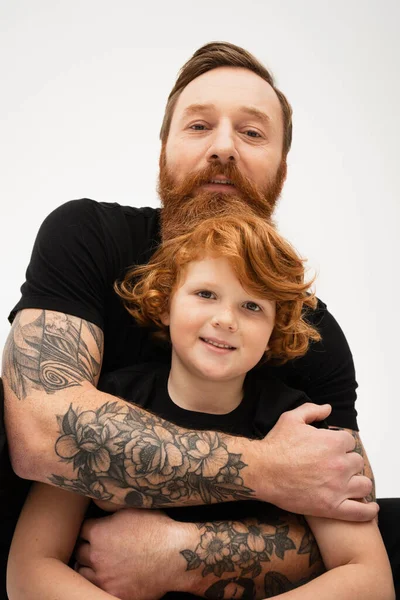 tattooed man in black t-shirt hugging red haired grandson smiling at camera isolated on grey