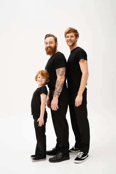 stock image full length of bearded redhead men and kid in black t-shirts and trousers smiling at camera while standing on light grey background