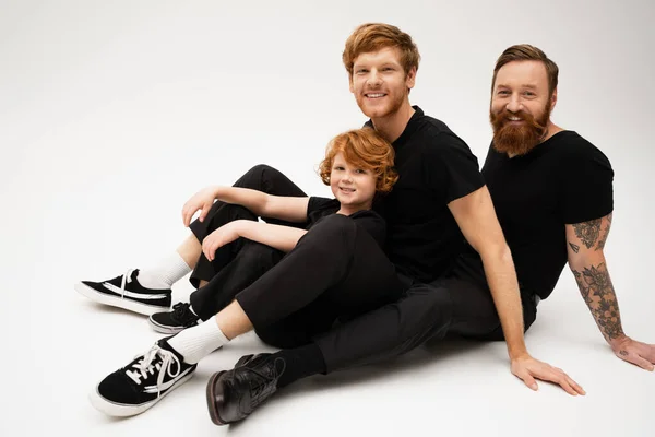 bearded tattooed man with redhead son and grandson smiling at camera while sitting on light grey background