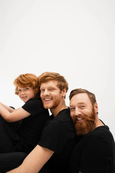 happy bearded man with redhead son and grandson in black t-shirts smiling at camera isolated on grey