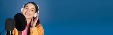 Cheerful teenager in headphones looking at camera near blurred studio microphone isolated on blue, banner 