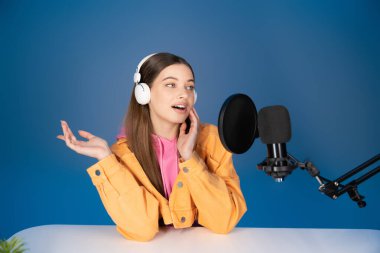 Teen girl in headphones talking during podcast near studio microphone isolated on blue 