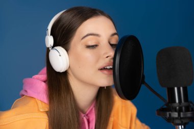 Brunette teenager in headphones talking during podcast isolated on blue  clipart