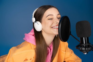 Cheerful teenager in headphones talking near studio microphone during podcast isolated on blue 