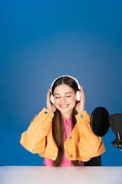 Cheerful brunette teenager in headphones sitting near microphone and table isolated on blue 