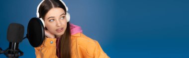 Teen girl in wireless headphones looking away near studio microphone isolated on blue, banner 