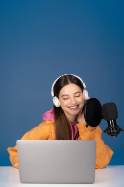 Smiling teenage girl in headphones looking at laptop near studio microphone isolated on blue  clipart