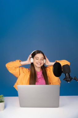 Smiling teen girl in headphones using laptop near microphone isolated on blue 