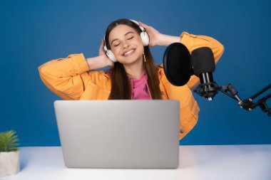 Happy teenager touching headphones near laptop and studio microphone isolated on blue 