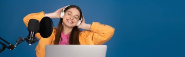 Teenager with closed eyes touching headphones near laptop and studio microphone isolated on blue, banner 