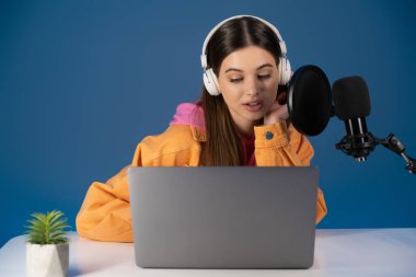Brunette teenager in headphones talking near studio microphone and laptop on table isolated on blue 