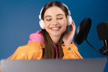 Pleased teenager in headphones looking at laptop near microphone isolated on blue 