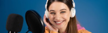 Teen podcaster in headphones smiling at camera near studio microphone isolated on blue, banner 