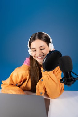 Positive teenage podcaster in headphones using laptop and studio microphone isolated on blue 