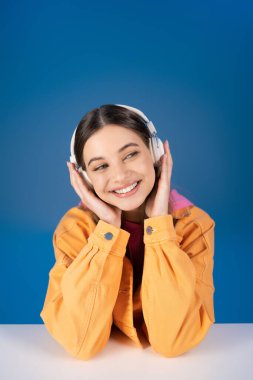 Positive teenager in headphones looking away near table isolated on blue 