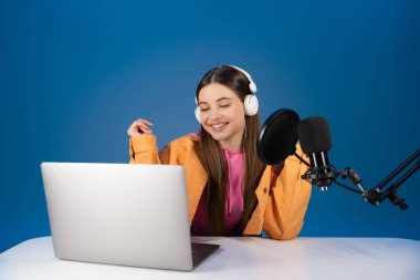 Positive teenager in headphones recording podcast near laptop and microphone isolated on blue 