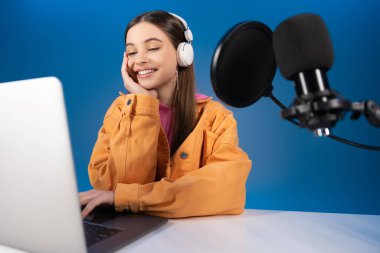 Teenager in headphones doing podcast near laptop and microphone isolated on blue 