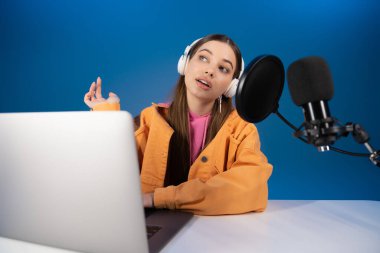 Teen girl in headphones talking in microphone during podcast isolated on blue 