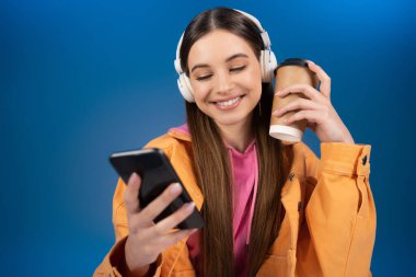 Positive teenager in headphones using cellphone and holding coffee to go isolated on blue 