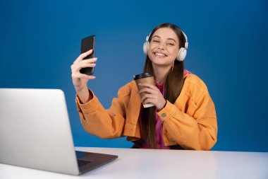 Positive teenager in headphones holding smartphone and coffee near laptop on table isolated on blue 